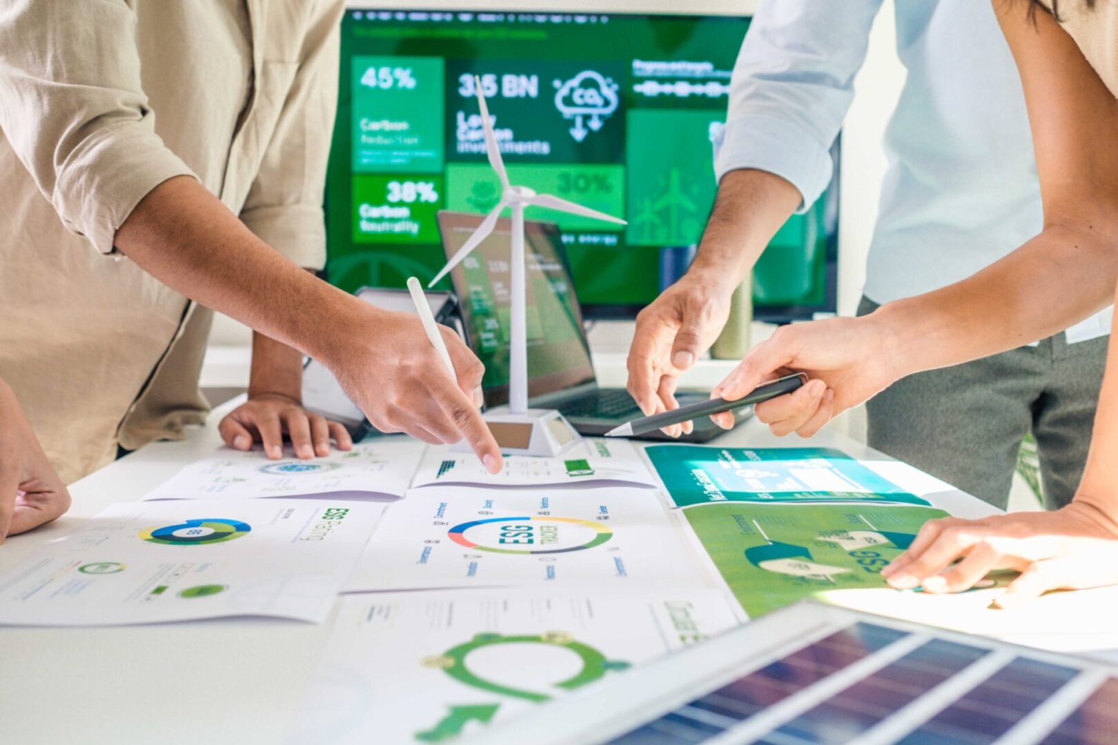jpeg-optimizer_GettyImages-2019990812 Our mission brings together a group examining documents on environmental data and renewable energy, with a model wind turbine and digital screens displaying statistics in the background.